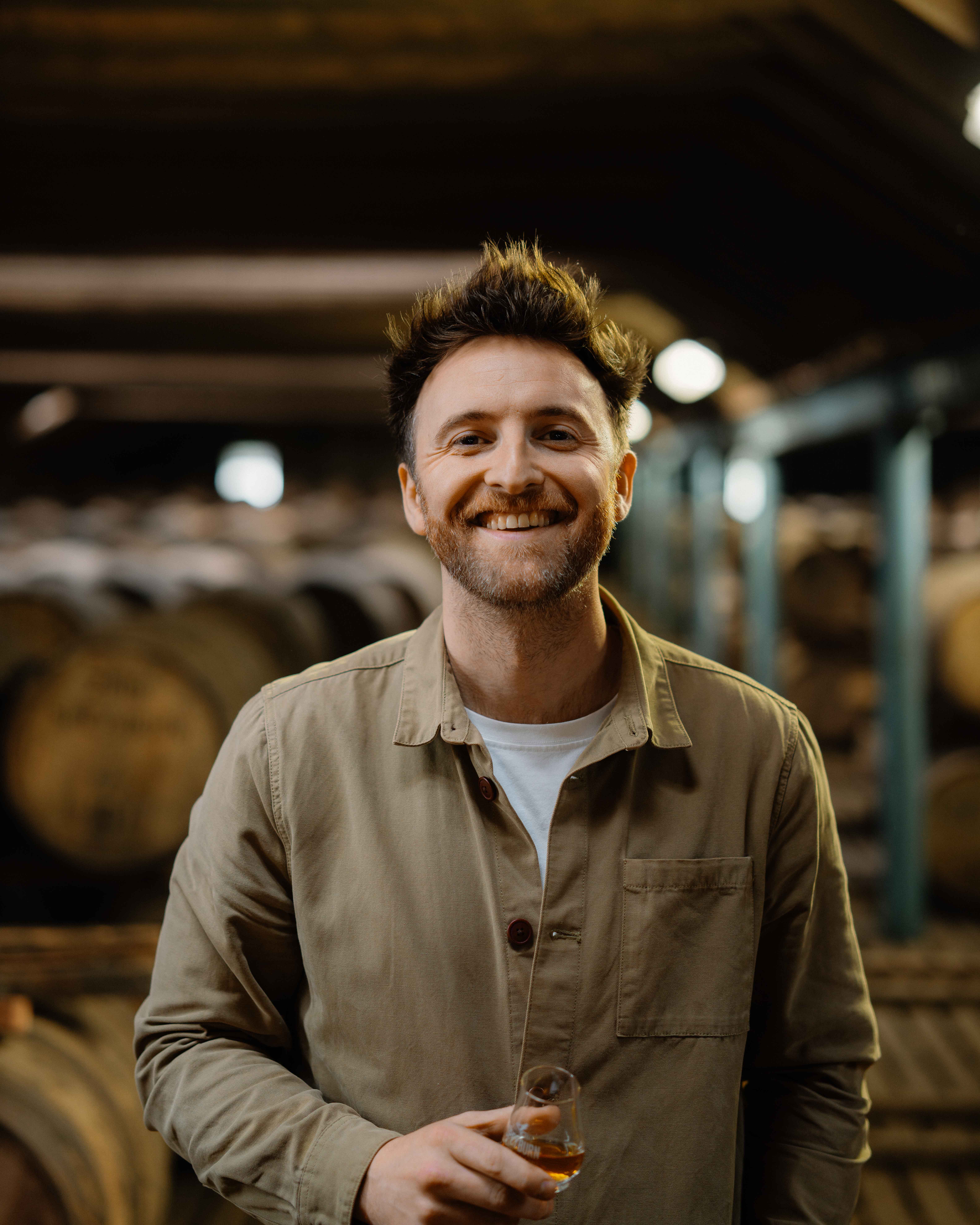 A man in a brown shirt stands in from of a number of wooden casks. He is holding a glass of whisky